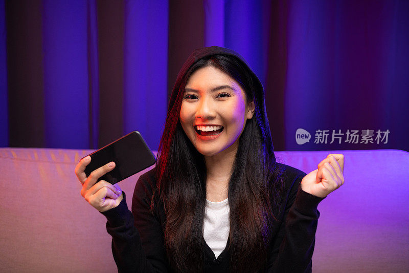 用手机玩游戏。年轻的亚洲漂亮女人坐在沙发上拿着智能手机在客厅。快乐女性Pro Streamer中国人穿着连帽衫玩游戏与霓虹灯。